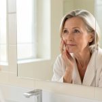 Femme de 50 ans appliquant une crème visage légère devant un miroir dans une salle de bain lumineuse, avec une peau naturelle et une ambiance douce.